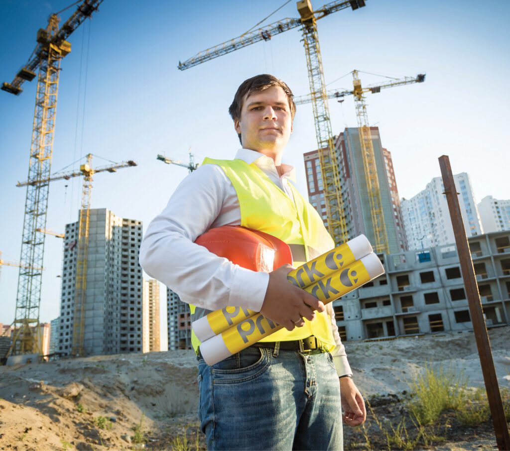 A contractor standing in an open job site wearing a yellow safety vest. He is holding his orange hard hat and 2 pipes with yellow coiled pipe markers that read premark in Gray. In the background, there are skyscrapers under construction with a clear view of four cranes.