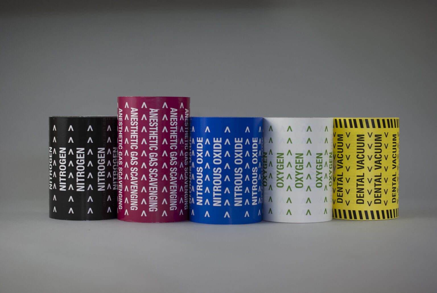 Five different medical pipe markers coiled up into rolls are standing vertically, showcasing the different colors and labels. From left to right, the colors go: black red, blue, white, and yellow