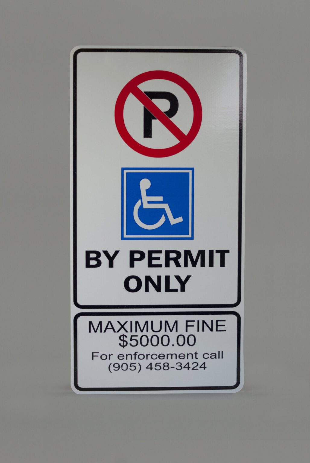 A white no parking sign that has a blue accessibility wheelchair icon with the text below that reads by permit only. Below that Explains the fines and Penalties