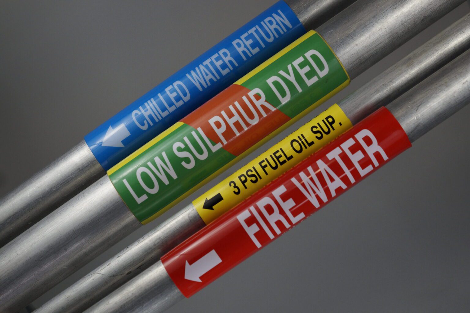 Four metal pipes with various widths showcasing four different coiled conduit markers showcasing their different colors and varieties on a 45° angle. The first coiled conduit marker is blue, and the legend says , Chilled water return with an arrow to the right pointing down. The second one is a yellow coiled pipe marker wrapped around a steel pipe. There is a green band going through the middle and a red stripe going through as a diagonal. The legends on the top and bottom read diesel, and the legend in Plainview reads. Low sulphur dyed. The third one is thinner and yellow with the legend reading 3 PSI Oil sup. The lastThe last pipe has a red coiled conduit marker that reads fire water.