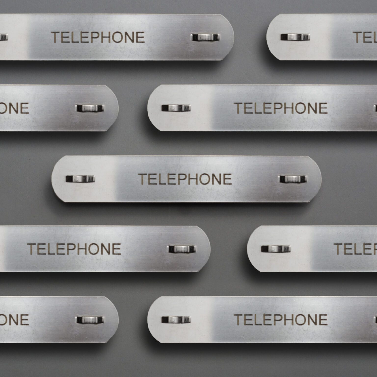 A display of long thin stainless steel conduit tags that read telephone are displayed nicely against a clean gray background.