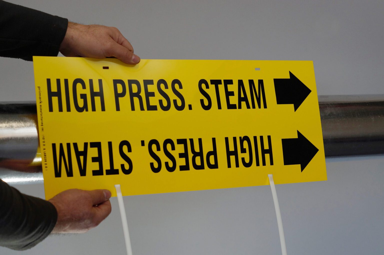 A man demonstrates how to apply the tie strap marker to a metal pipe. The man places the marker flat in front of the pipe with the zip ties facing down demonstrating how to apply. The marker is yellow with black text legend reads high pressure steam with an that points to the right