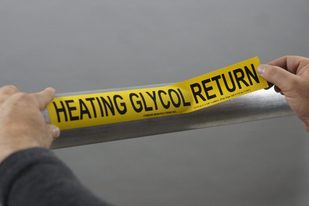 A close up of a man applying an adhesive pipe marker to a steel pipe. One hand secures the sticker to the pipe while the other hand is holding the sticker that has not been applied yet. The marker is yellow and the legend reads "Heating glycol return"