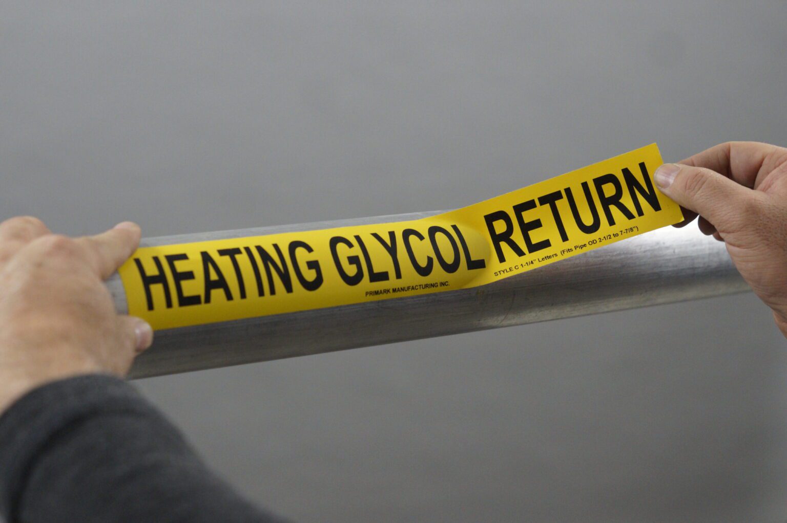 A close up of a man applying an adhesive pipe marker to a steel pipe. One hand secures the sticker to the pipe while the other hand is holding the sticker that has not been applied yet. The marker is yellow and the legend reads "Heating glycol return"
