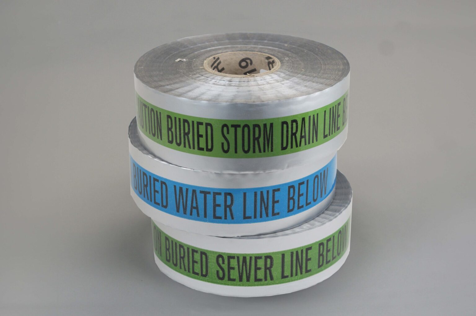 A stack of three detectable underground tape rolls. The bottom one is green the middle one is blue and the top one is also green there is a silver reflective coating on all of them