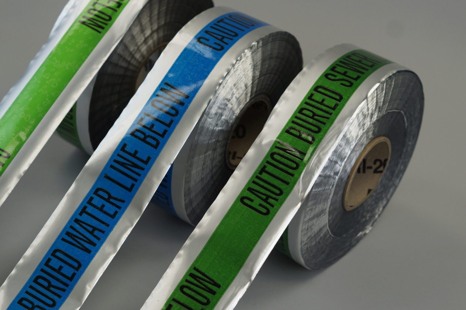 Three detectable underground tape rolls are standing vertically with their tape stretched out to the ground. The order from left to right goes green blue green and there is a silver reflective layer on all of them.