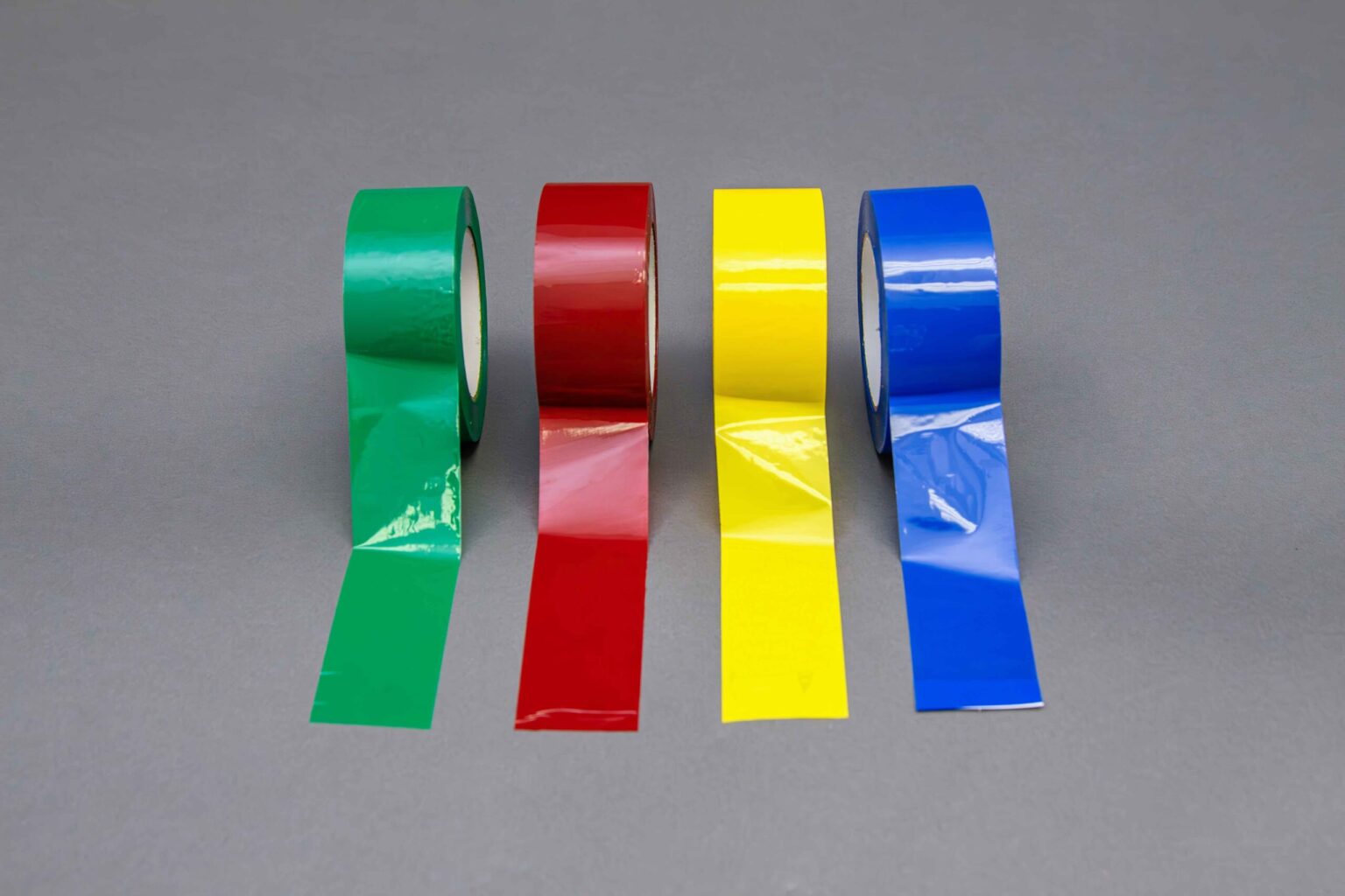 Four different colored banding tapes sit side by side with a portion of the tape extended onto the clean gray background. The first is blue, the second is yellow, the third is red, and the last is green. Looking straight on