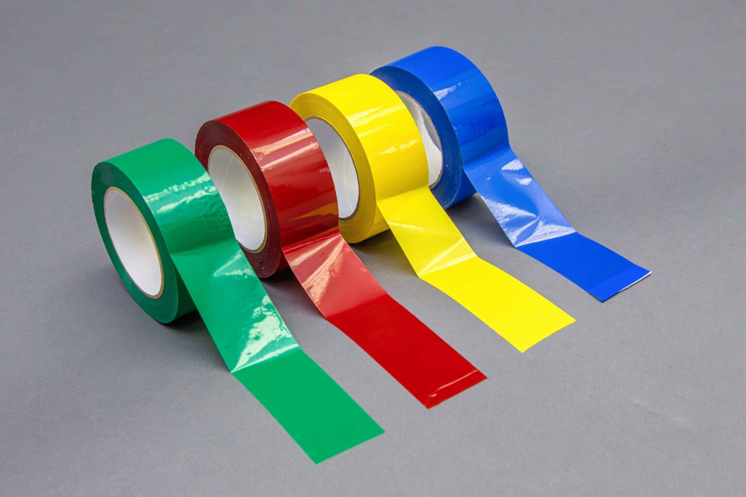 Four different colored banding tapes sit side by side with a portion of the tape extended onto the clean gray background. The first is blue, the second is yellow, the third is red, and the last is green. On a 45° angle