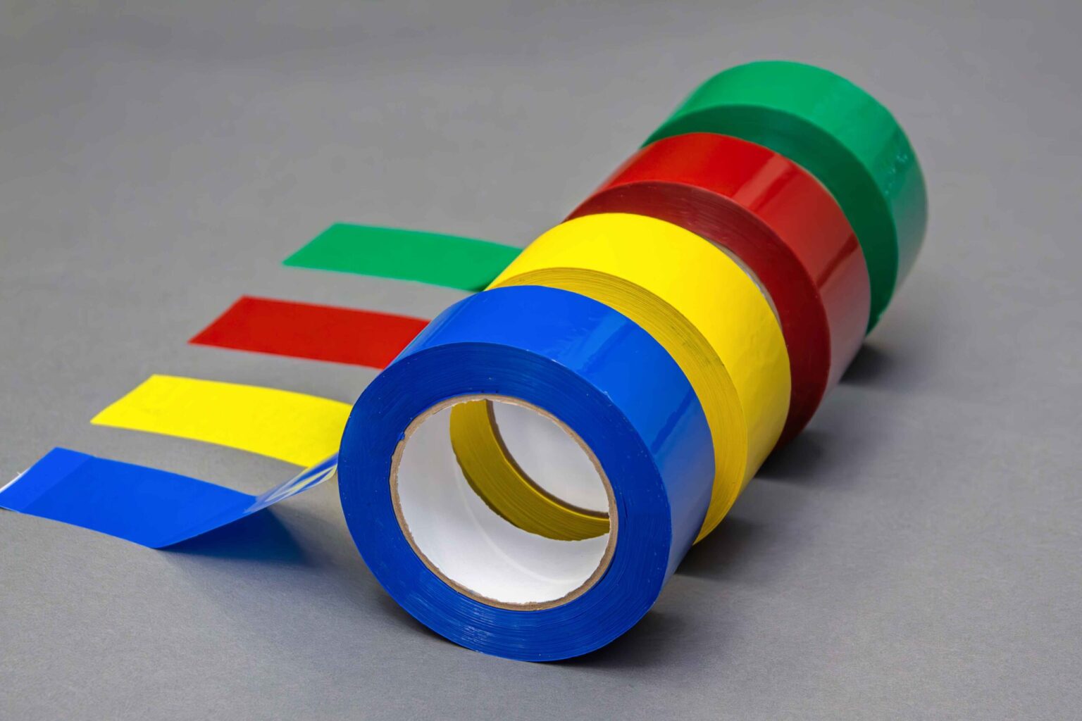 Four different colored banding tapes sit side by side with a portion of the tape extended onto the clean gray background. The first is blue, the second is yellow, the third is red, and the last is green.