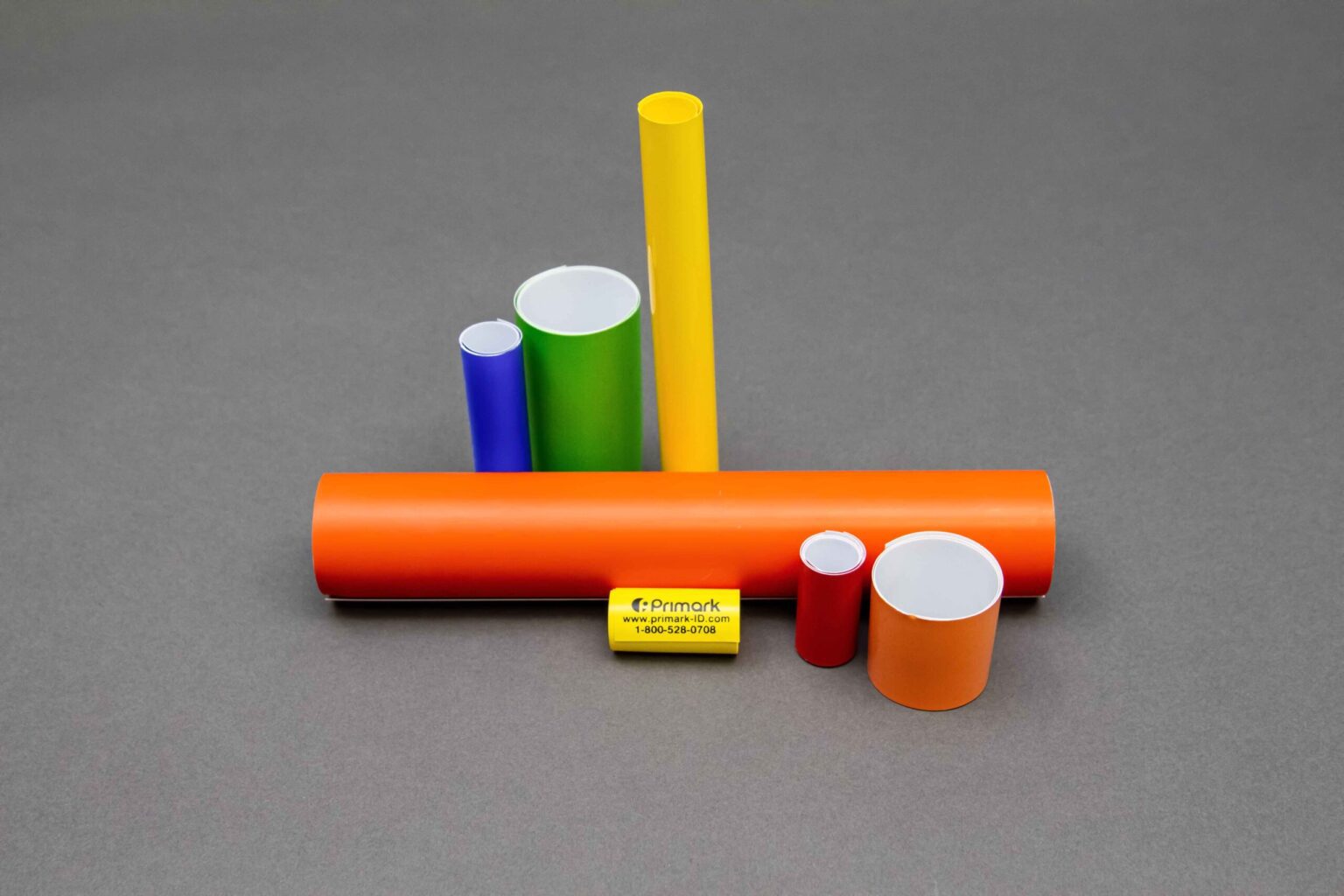 7 coiled conduit markers are displayed nicely against a clean gray background. To the left there are two small coils one green one blue a tall yellow Coil In The Middle all Standing vertically. In front is a long orange coiled conduit marker sitting horizontal in the middle, to the left is a short red and orange Coiled conduit marker and in the very front is a small yellow conduit marker with the Primark logo address and phone number