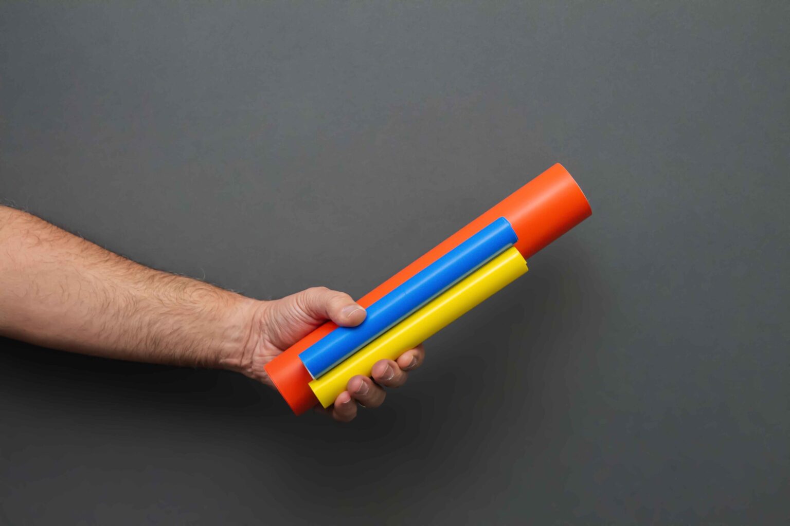 A hand is holding three different coiled conduit markers Two small ones and one big one. The two small ones are blue and yellow and the tall big one is orange against a dark gray background