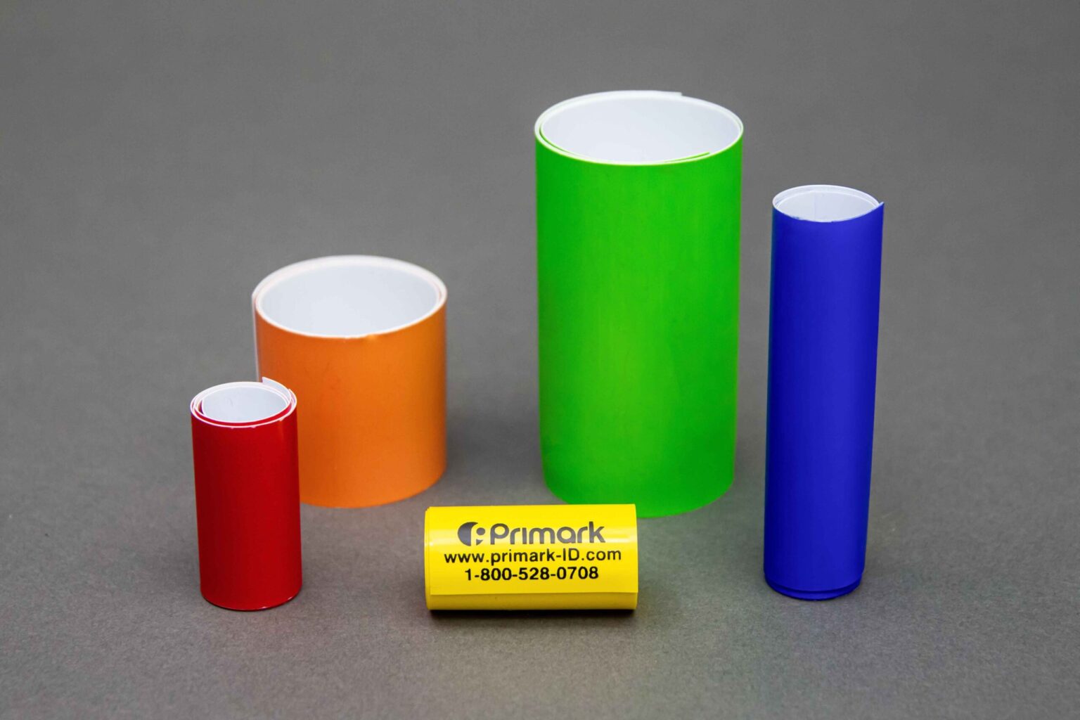 Five coiled conduit markers of different sizes and colors are displayed nicely on a clean gray background. The four bigger ones are standing vertically. The first and the smallest standing vertically is short in red to the left is a thicker orange one but not as tall to the left of that is a taller green one that is thicker and to the left of that is a thin blue coiled conduit marker. In the front sitting horizontally is a very small yellow marker with the Primark logo address and phone number on it