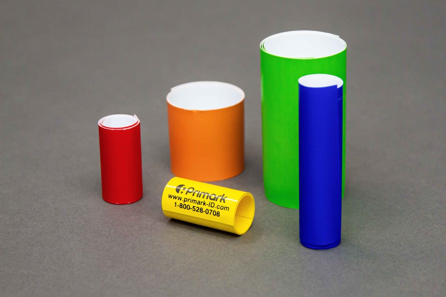 Five coiled conduit markers of different sizes and colors are displayed nicely on a clean gray background. The four bigger ones are standing vertically. The first and the smallest standing vertically is short in red to the left is a thicker orange one but not as tall to the left of that is a taller green one that is thicker and to the left of that is a thin blue coiled conduit marker. In the front sitting horizontally is a very small yellow marker with the Primark logo address and phone number on it