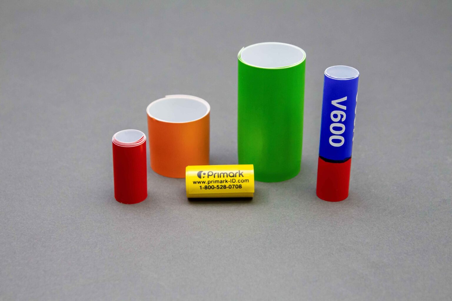 Five coiled conduit markers of different sizes and colors are displayed nicely on a clean gray background. The four bigger ones are standing vertically. The first and the smallest standing vertically is short in red to the left is a thicker orange one but not as tall to the left of that is a taller green one that is thicker and to the left of that is a thin blue coiled conduit marker. In the front sitting horizontally is a very small yellow marker with the Primark logo address and phone number on it
