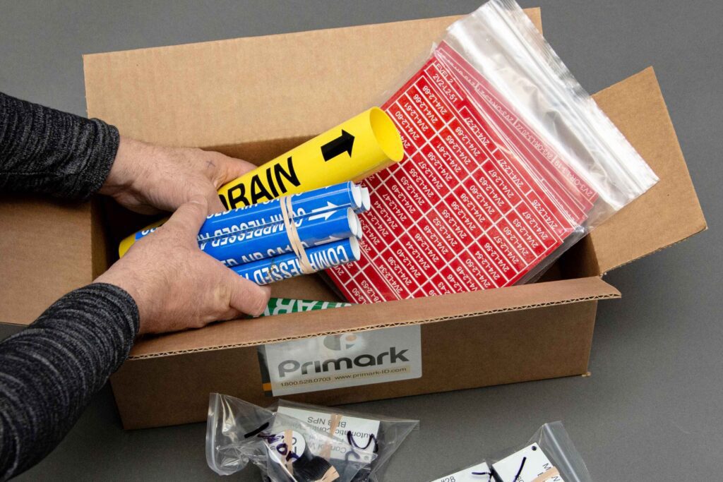 A man loads coiled pipe markers, tags, and engraved labels into an open brown box with the Primark logo on the side. The box is on a clean gray floo