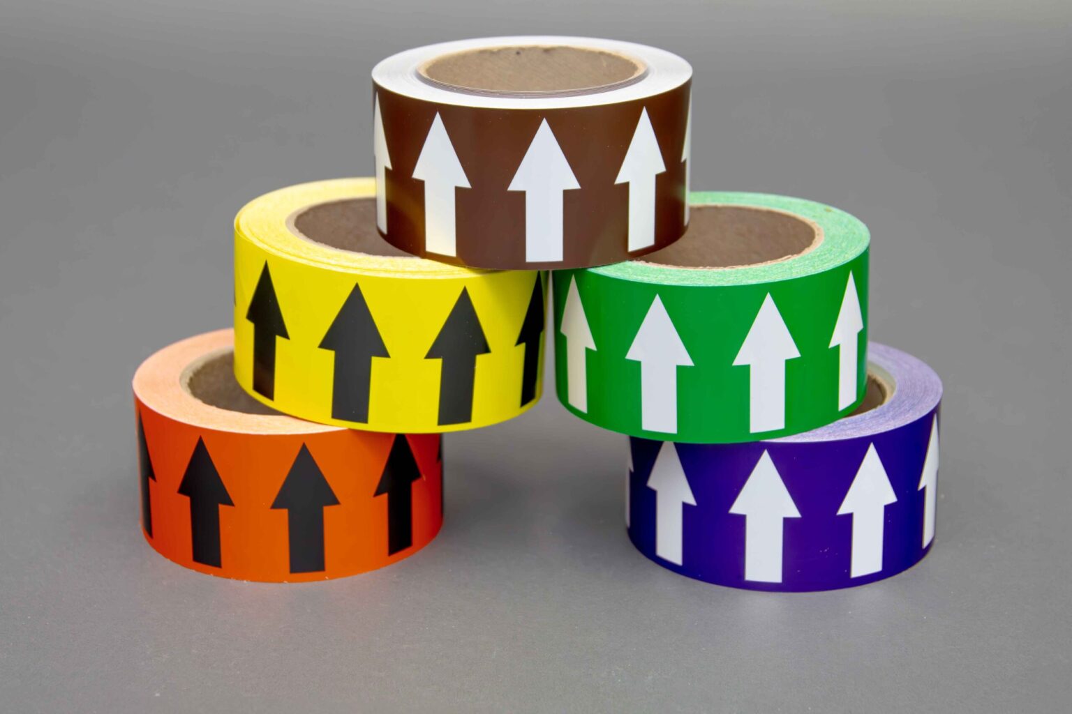 Five different colored arrow taps are stacked on top of each other to form a triangle with all of the arrows pointing up. The bottom has an orange roll with black arrows spread out from a purple roll with white arrows. Above that is a green roll with white arrows and a yellow roll with black arrows. Sitting at the top is a Brown tape roll. Displayed nicely against a clean gray background