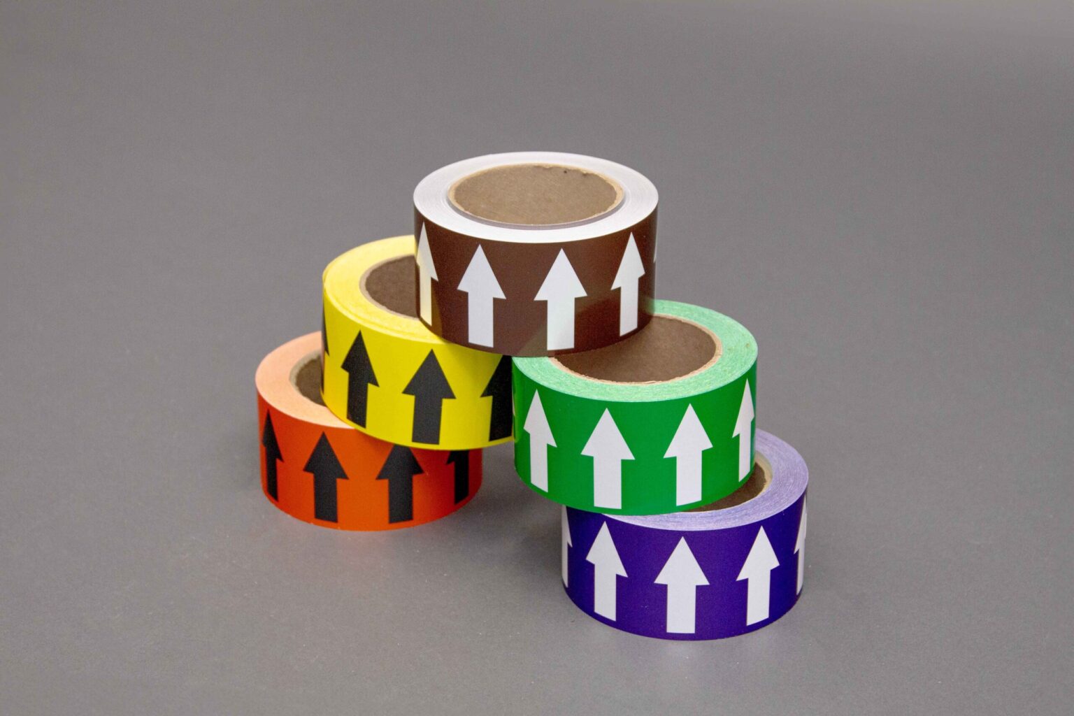 Five different colored arrow taps are stacked on top of each other to form a triangle with all of the arrows pointing up. The bottom has an orange roll with black arrows spread out from a purple roll with white arrows. Above that is a green roll with white arrows and a yellow roll with black arrows. Sitting at the top is a Brown tape roll. Displayed nicely against a clean gray background on a 45° angle