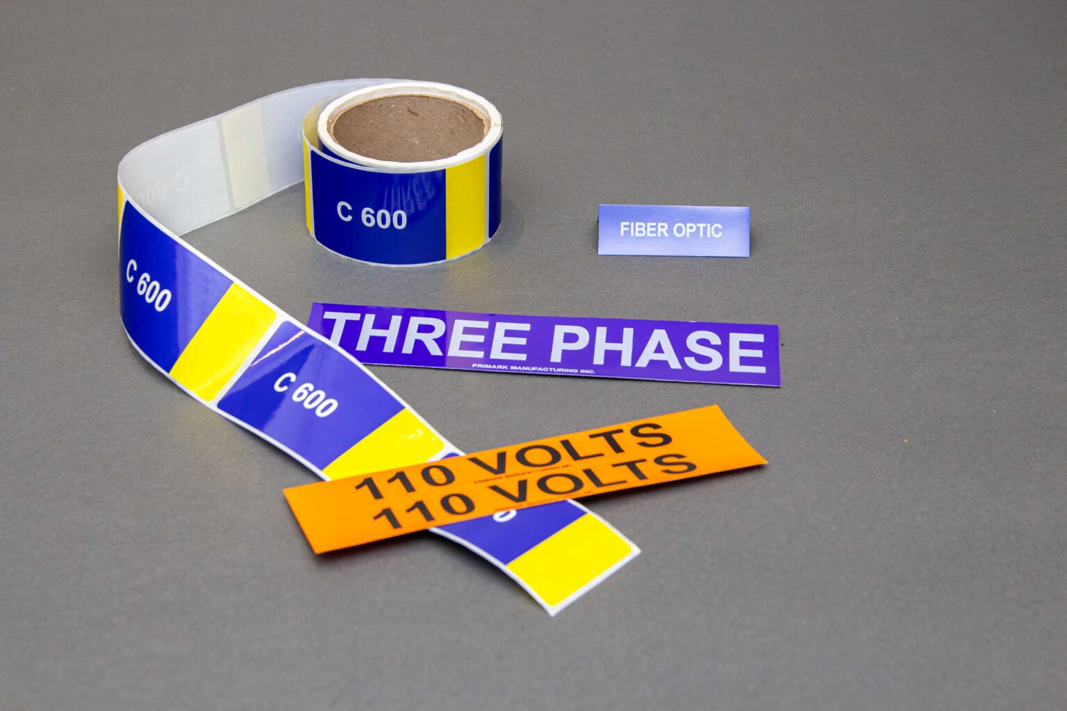 There is an intricate Gathering of four different types of adhesive conduit markers. A roll of blue and yellow stickers labeled c600 there is also a purple sticker that reads three phase, an orange sticker that reads 110 volts and a small blue sticker that reads fiber optic on a clean gray background
