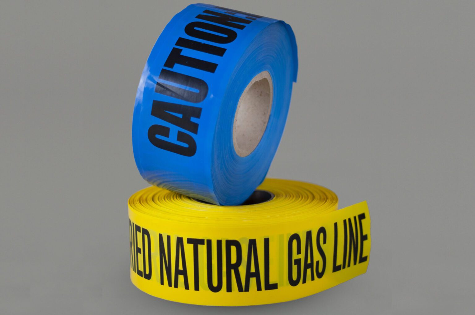 At the bottom, there is a roll of Non-Detectable underground tape that reads gas line below. Stacked on top is a blue non-detectable underground tape roll that reads caution. On a clean white background
