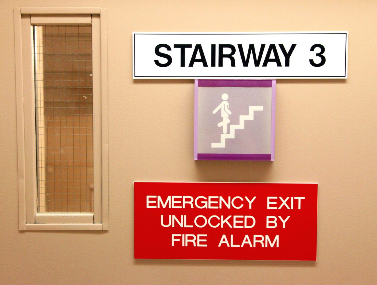 A close up of a industrial stairwell with the door closed focusing on the emergency exit sign that reads "emergency exit unlocked by fire alarm"