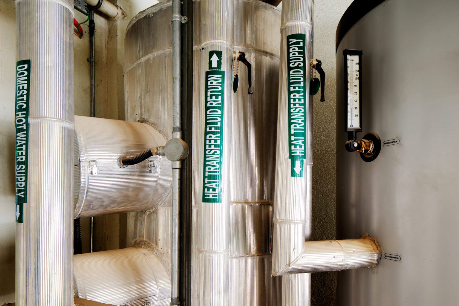 A busy boiler room with three vertical pipes that have green adhesive pipe markers applied to them. The first Legend reads domestic hot water supply with an arrow that points down. The second pipe marker Legend reads "heat transfer fluid return" with the arrow facing up. The last pipe reads "heat transfer fluid Supply" with an arrow pointing down