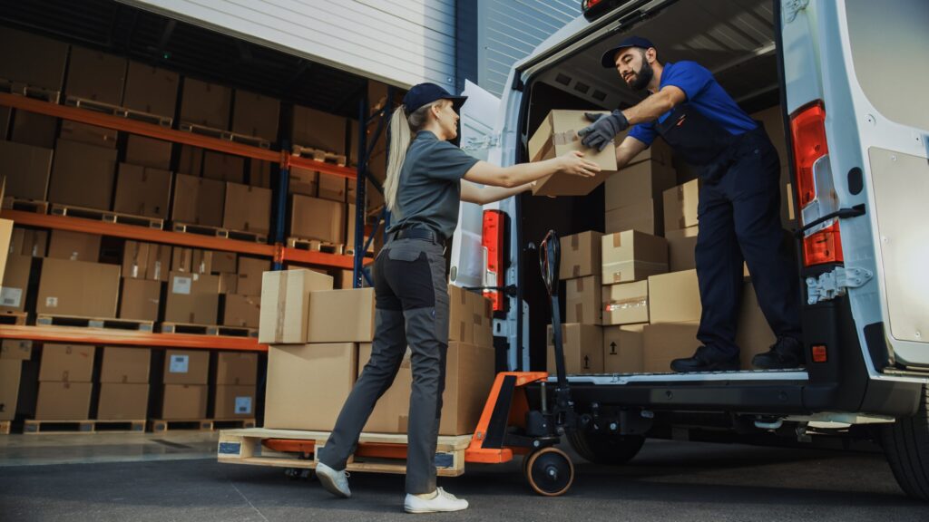 A delivery truck full of boxes is parked in front of a warehouse also full of boxes. The back door is open to a delivery man receiving a package from a delivery woman on the ground. There is a cart full of boxes beside her.