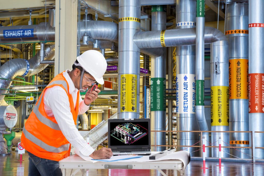 An engineer using a walkie-talkie in the maintenance room of a building showcasing the different mechanical markers on each pipe