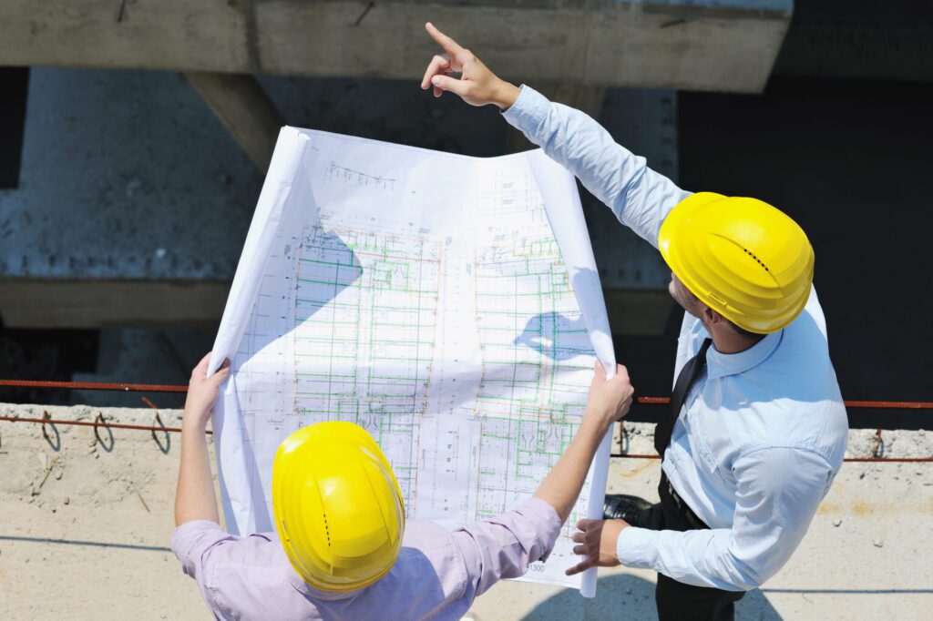 A overhead view of two contractors on a job site with yellow hard hats. One of them is holding a blueprint of the structure the other is pointing off in the distance