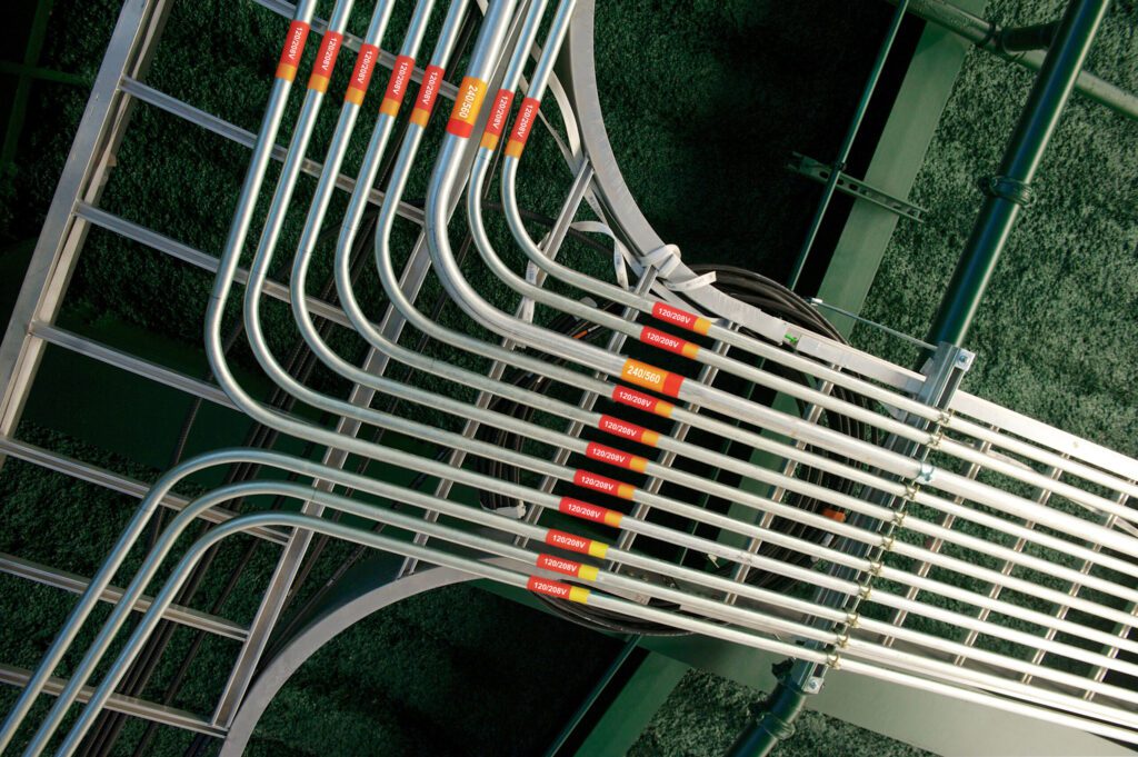 A industrial ceiling with an intricate 11 conduit ceiling with red and orange coiled conduit markers indicating the voltage of each conduit