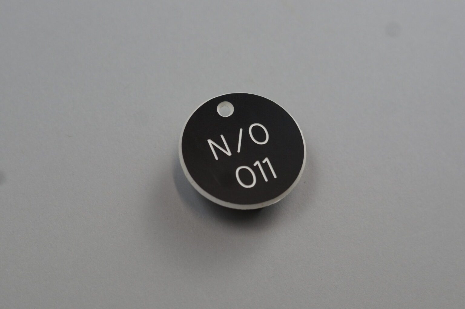 A circular black valve tag with a hole at the top white text reads "N / O 011" against the clean gray background
